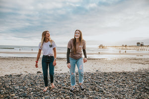 Two girls smiling and laughing on the beach by the ocean wearing reef safe orange and blue sunscreen on nose